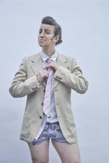 © Jana Rothe Drag King character Richard P. Dick Van Johnson wears a tan suit jacket, no pants, and a matching pink tie and underwear. He's fixing his tie.