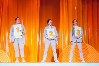 © Mayra Wallraff Drei Performerinnen in grauen Anzügen stehen vor einem orangefarbenen Vorhang und halten drei Tafeln mit den Nummer 1, 2 und 3 in den Händen.