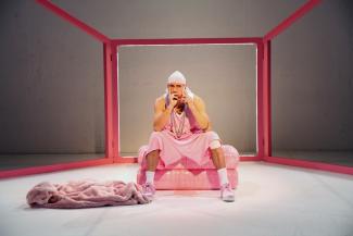© Dorothea Tuch A performer, dressed in a loose sleeveless jersey, pink sports pants and a white bandana, holds a cell phone to his ear with his right hand. He is sitting on a pink cushion. In front of him in the left half of the image is a pink plush garment on the floor. His playing surface is framed by a framework of pink struts.
