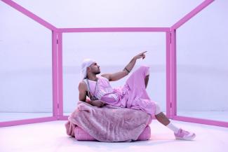 © Dorothea Tuch A performer, dressed in a loose sleeveless jersey, pink sports pants and a white bandana, lies on a pink cushion. His left index finger points into the distance, and his left leg is bent. The pose is modeled after Michelangelo's ceiling fresco, "The Creation of Adam." In the background, the stage set is framed by a framework of pink struts.