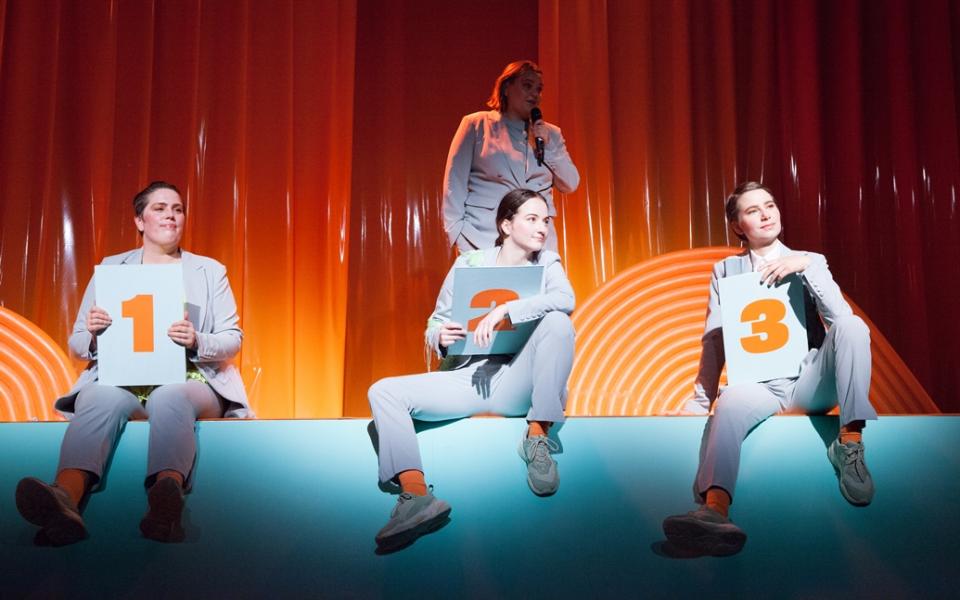 Drei Performerinnen in grauen Anzügen sitzen auf einem Bühnenrand und halten drei Tafeln mit den Nummer 1, 2 und 3 in den Händen. Im Hintergrund steht eine Frau vor einem orangefarbenen Vorhang und spricht in ein Mikro. 