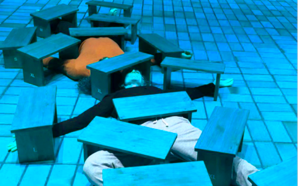 Zwei Körper liegen unter vielen Holzbänkchen, so dass nur ein Oberkörper in orange sowie Beine in beige erkennbar sind. Der Boden ist von Kacheln durchzogen und das Bild schimmert in einem blauton. s