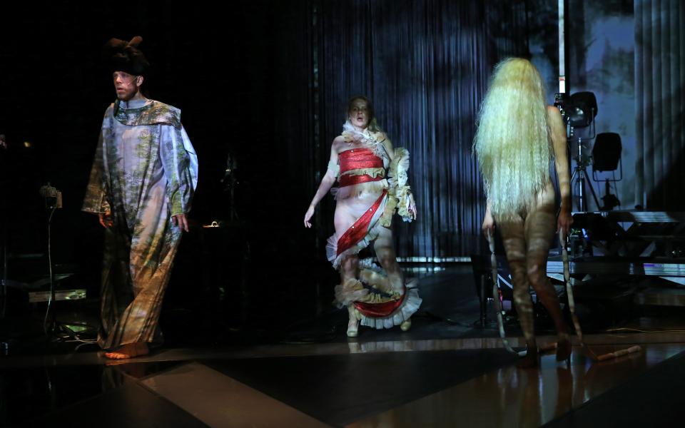 Zwei Performer:innen von She She Pop stehen in Kostümen auf der dunklen Bühne. Eine Performerin ist nackt, ihr Körper wird von einer blonden Langhaarperücke bedeckt.