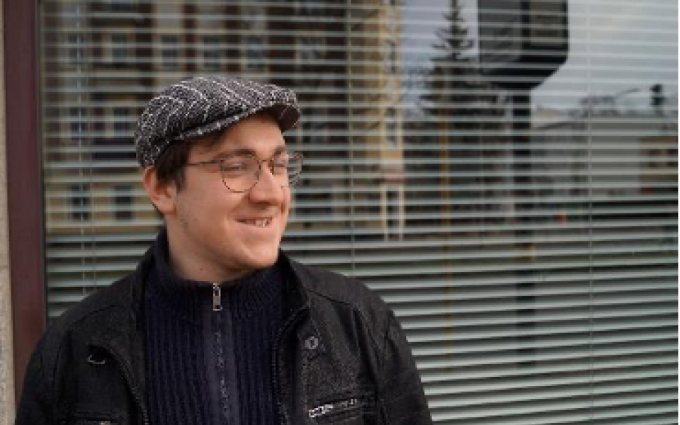 Marius Zoschke Porträt von Marius Zoschke mit schwarzer Jacke und Schiebermütze, hinter ihm ein Fenster, darin spiegelt sich die Stadt
