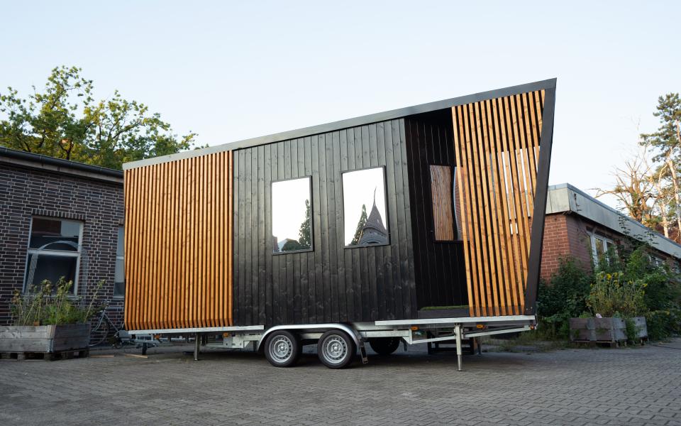 A Tiny House on casters clad in black and teak-stained wood slats stands in an undefined outdoor space. The windows are mirrored.