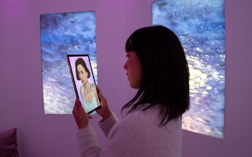 A person, probably read as female, stands in an interior room in front of two screens. She holds a tablet that shows a humanoid avatar.  The indirect room light colors the walls purple-pink