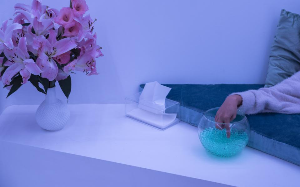 A hand reaches from outside the picture frame into a glass bowl with jelly beads. Next to it is a handkerchief dispenser and a bouquet of flowers. The indirect room light colors the walls ice blue. 
