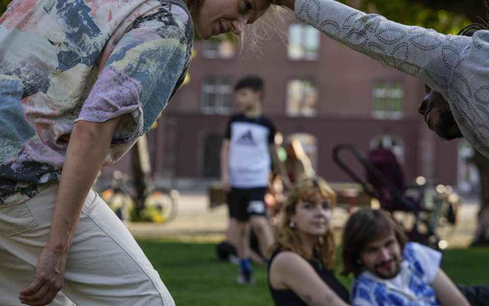 Melissa ist eine weiße Frau mit langen blonden Haaren, die zu einem Pferdeschwanz gebunden sind. Sie trägt ein buntes Hemd und eine weiße Hose. Sie lächelt, während sie ihre Knie beugt und ihren Kopf und ihr Gesicht nach vorne zu einem anderen Tänzer neigt, der sie anlächelt und mit seiner ausgestreckten rechten Hand ihren Kopf hält. Sie befinden sich in einem Park mit grünem Gras und einem grünen, üppigen Baum im Hintergrund sowie einem Publikum, das aus der Nähe zuschaut.