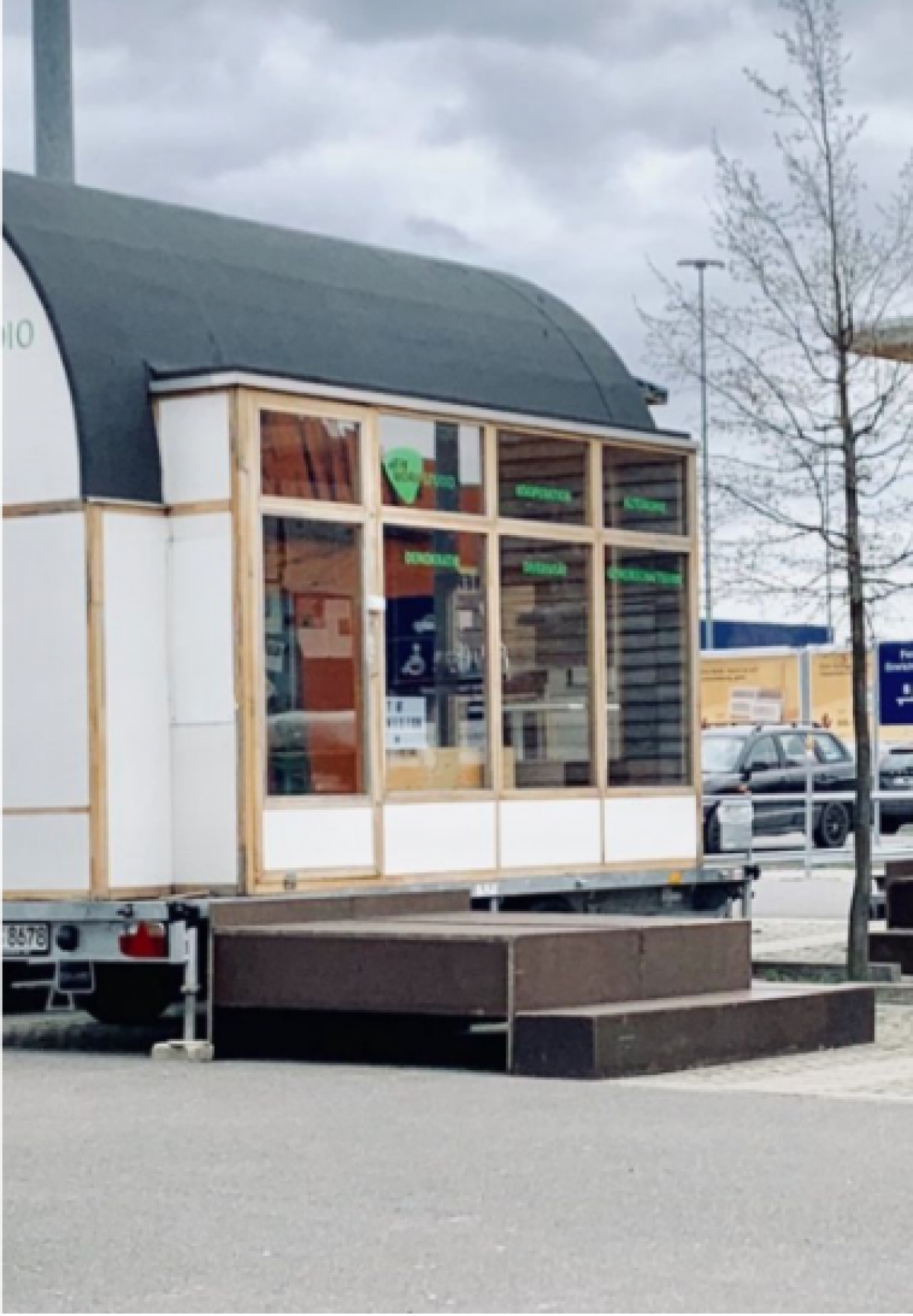 Tiny House Ein kleines Häuschen mit abgerundetem grauen Dach und einer Glasfront. Davor steht ein kleines Podest.