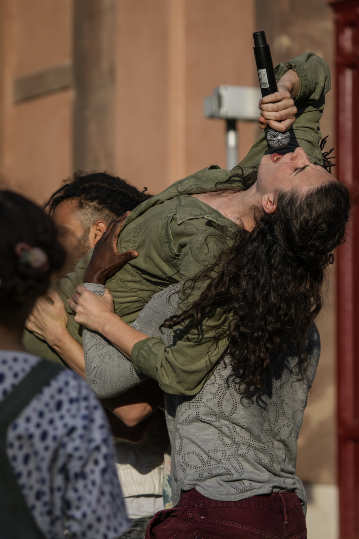 © Matthias Wäckerlin Katy is a white woman with long brown hair that falls down her back. She is wearing an olive green long-sleeved jumpsuit. Her head is tilted back and she sings energetically into a microphone she holds in her right hand while being carried on her shoulders by three other dancers. The wall of a sandstone-coloured house can be seen in the background.