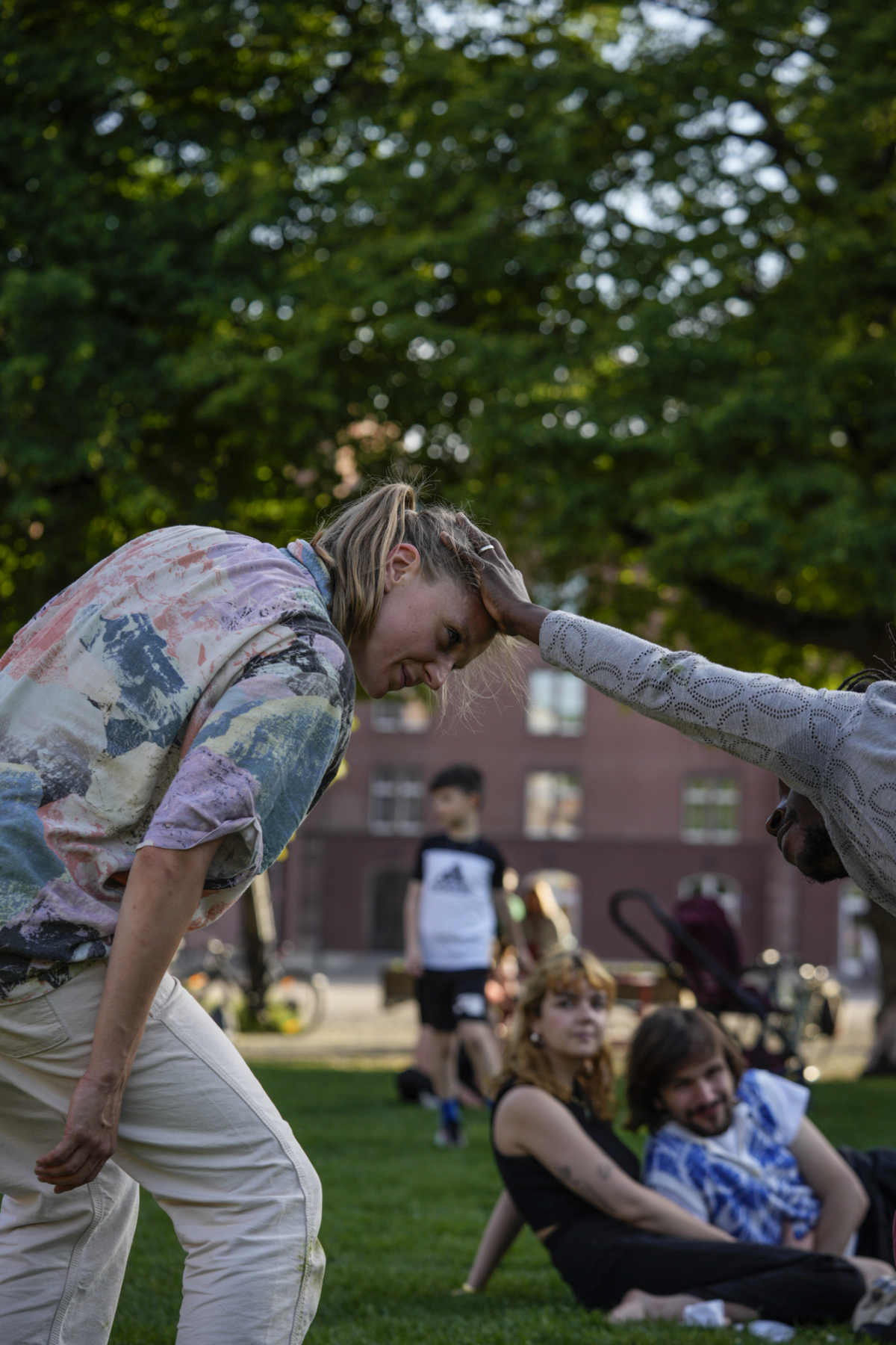 © Matthias Wäckerlin Melissa is a white woman with long blonde hair tied back in a ponytail. She is wearing a colourful shirt and white trousers. She smiles as she bends her knees and tilts her head and face forward towards another dancer who smiles at her and holds her head with his outstretched right hand. They are in a park with green grass and a green lush tree in the background and an audience watching from close by.