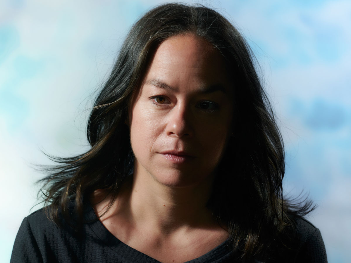 © Charlotte Krieger Portrait photo of Mirjam Gurtner against a light blue cloudy background. A white woman, with brown eyes and brown long hair worn loose, and a dark blue T-shirt.
