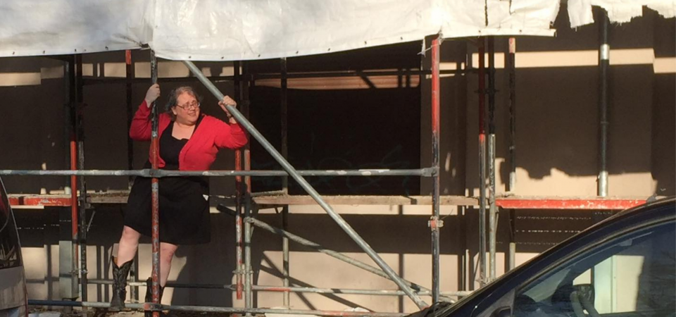 Cameryn Moore | Street Dog Stories A white woman wearing a short black dress, coral cardigan sweater, black cowboy boots, and glasses is climbing up some scaffolding in front of a building. She is looking out to her left and smiling slightly, or maybe she is squinting in the afternoon soon. The edge of a white tattered tarp flaps behind her.