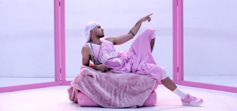 Ein Performer, der bekleidet ist mit einem weiten ärmellosen Trikot, einer rosafarbenen Sporthose und einem weißen Bandana, liegt auf einem rosafarbenen Polster. Sein linker Zeigefinger zeigt in die Ferne, sein linkes Bein ist angewinkelt. Die Pose ist Michelangelos Deckenfresko "Die Erschaffung Adams" nachempfunden. Im Hintergrund wird das Bühnenbild umrahmt von einem Gerüst aus rosafarbenen Streben. 