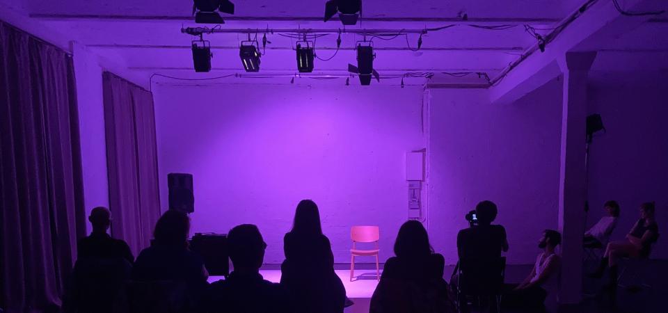 Shot of a stage illuminated in purple. The stage, which can be seen behind the audience, is empty.